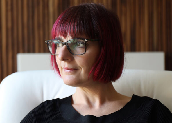 A person sitting on a white chair. The person has short red hair, is wearing glasses and a black top. The background consists of a wooden wall with vertical lines.