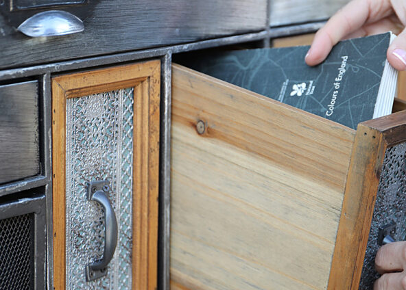 A hand pulling out a drawer from a wooden cabinet. The cabinet has multiple drawers with different designs and textures. The drawer being pulled out is made of wood and has a colour chart with a dark grey cover being pulled out.