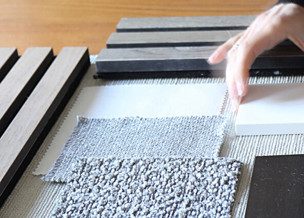 A person’s hand reaching for a fabric swatch on a table. The table is covered in various fabric and colour swatches. The swatches are arranged in a grid-like pattern. The person’s hand is reaching for a grey swatch with a textured pattern. The other swatches on the table are brown, white, and grey in colour and have different patterns and textures.