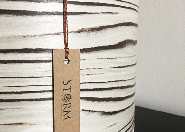 A light cappuccino cylindrical lampshade with brown horizontal stripes. The lampshade has a brown tag hanging from it with the word ‘STORM’ written on it in black. The tag is attached to the lampshade with a brown string. The background is a plain white wall.