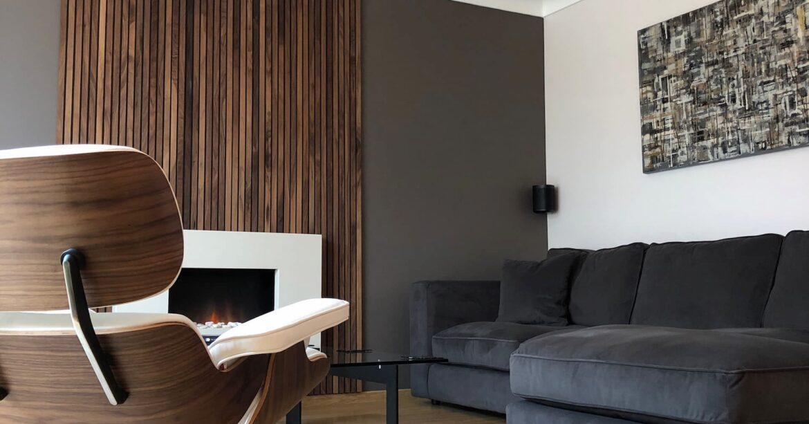 A modern living room with a fireplace and a wooden accent wall. The room has a wooden floor and a grey corner sofa with a throw and pillows. There is a white armchair with a wooden base and a coffee table with black legs and a glass top in front of the sofa. The fireplace is white and built into the wooden accent wall. There is a brown and white abstract painting hanging above the sofa. There is a decorative copper light pendant hanging from the ceiling.