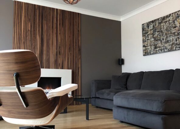 A modern living room with a fireplace and a wooden accent wall. The room has a wooden floor and a grey corner sofa with a throw and pillows. There is a white armchair with a wooden base and a coffee table with black legs and a glass top in front of the sofa. The fireplace is white and built into the wooden accent wall. There is a brown and white abstract painting hanging above the sofa. There is a decorative copper light pendant hanging from the ceiling.