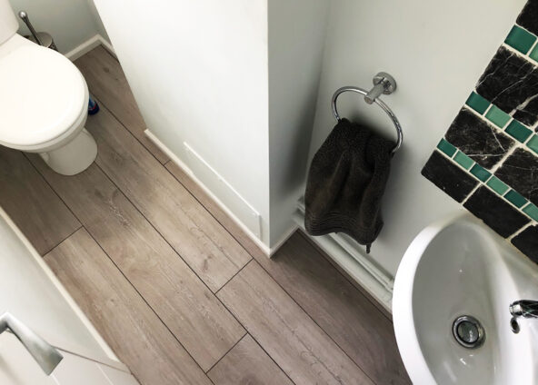 Image of a small downstairs cloakroom bathroom toilet, sink, and towel rail. The floor is made of grey wood-like tiles. The walls are white and there is a green and black tile backsplash behind the sink. The towel rail is silver and has a dark grey towel hanging on it. Photo has been taken from a high angle so it's a birds eye view.