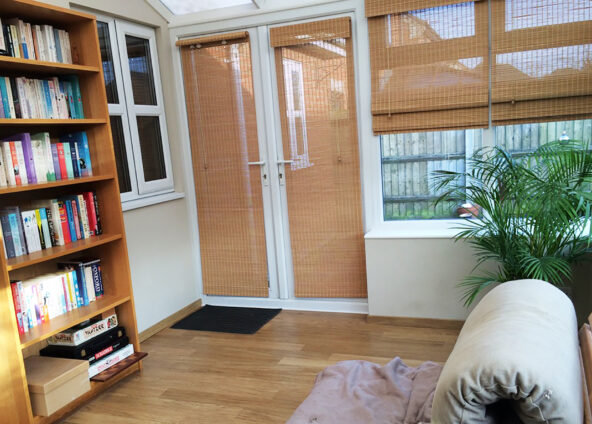 Showing the conservatory before the stained glass window was added. The bookcase is to the left of the window and there is a palm-type plant, and a futon opposite, there are bamboo blinds in front of the windows and doors.