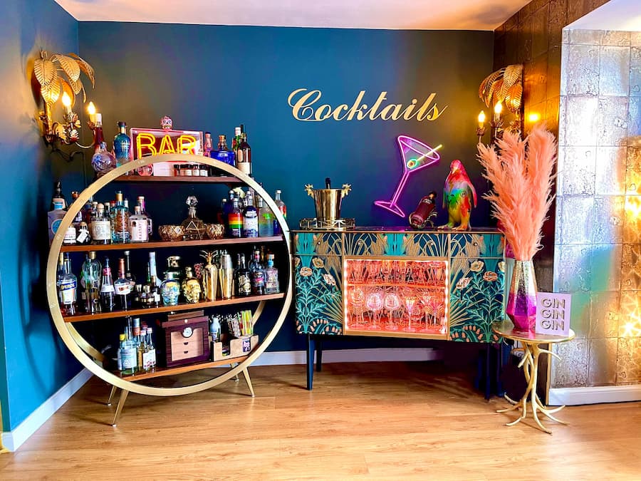 Bright vibrant lounge with cocktail area. Deep dark navy walls with gold cocktail sign on the wall. Circular drinks unit in gold to the left and decorative cabinet in turquoise and gold