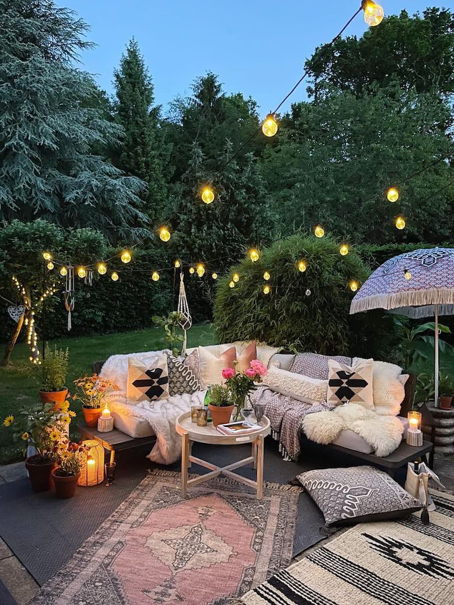 Outdoor living room with corner sofa covered in throws and cushions, an outdoor rug positioned in the centre and a parasol to the right of the sofa. Photoed at dusk with festoon lights