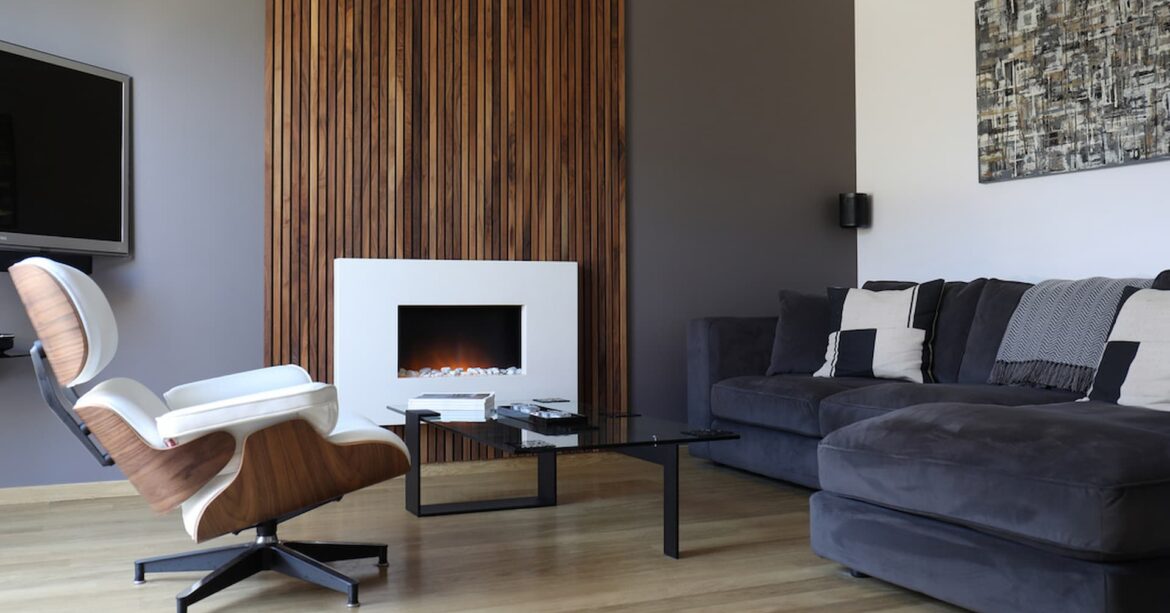 A modern living room with a fireplace and a wooden accent wall. The room has a wooden floor and a grey corner sofa with a throw and pillows. There is a white armchair with a wooden base and a coffee table with black legs and a glass top in front of the sofa. The fireplace is white and built into the wooden accent wall. There is a brown and white abstract painting hanging above the sofa.