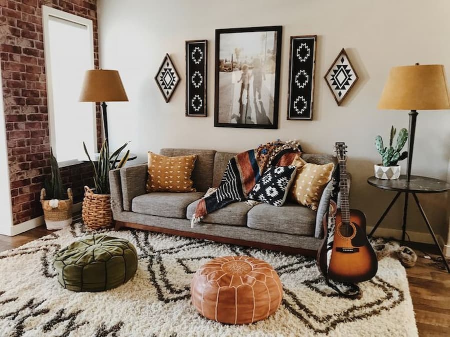 Bohemian decorated living room Three seater sofa in brown is positioned against the wall with aztec decorative through and cushions. There are tall lamps either side of the sofa with a yellow lampshade. In front of the sofa are two poufee cushions, one in olive green, one in tan leather which are sat on a white and black rug.