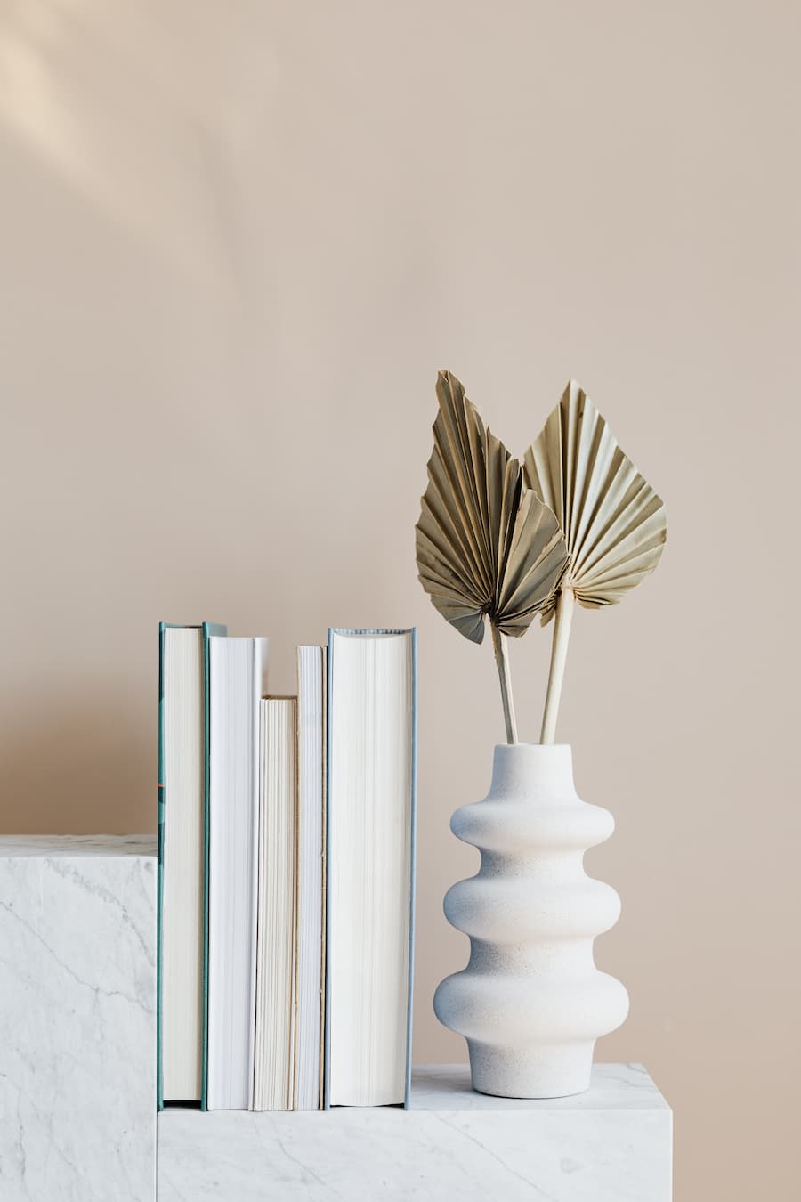 Simple set up of 5 books in varying widths showing the pages rather than the spine of the book alongside a white ribbed vase with two dried ferns inside