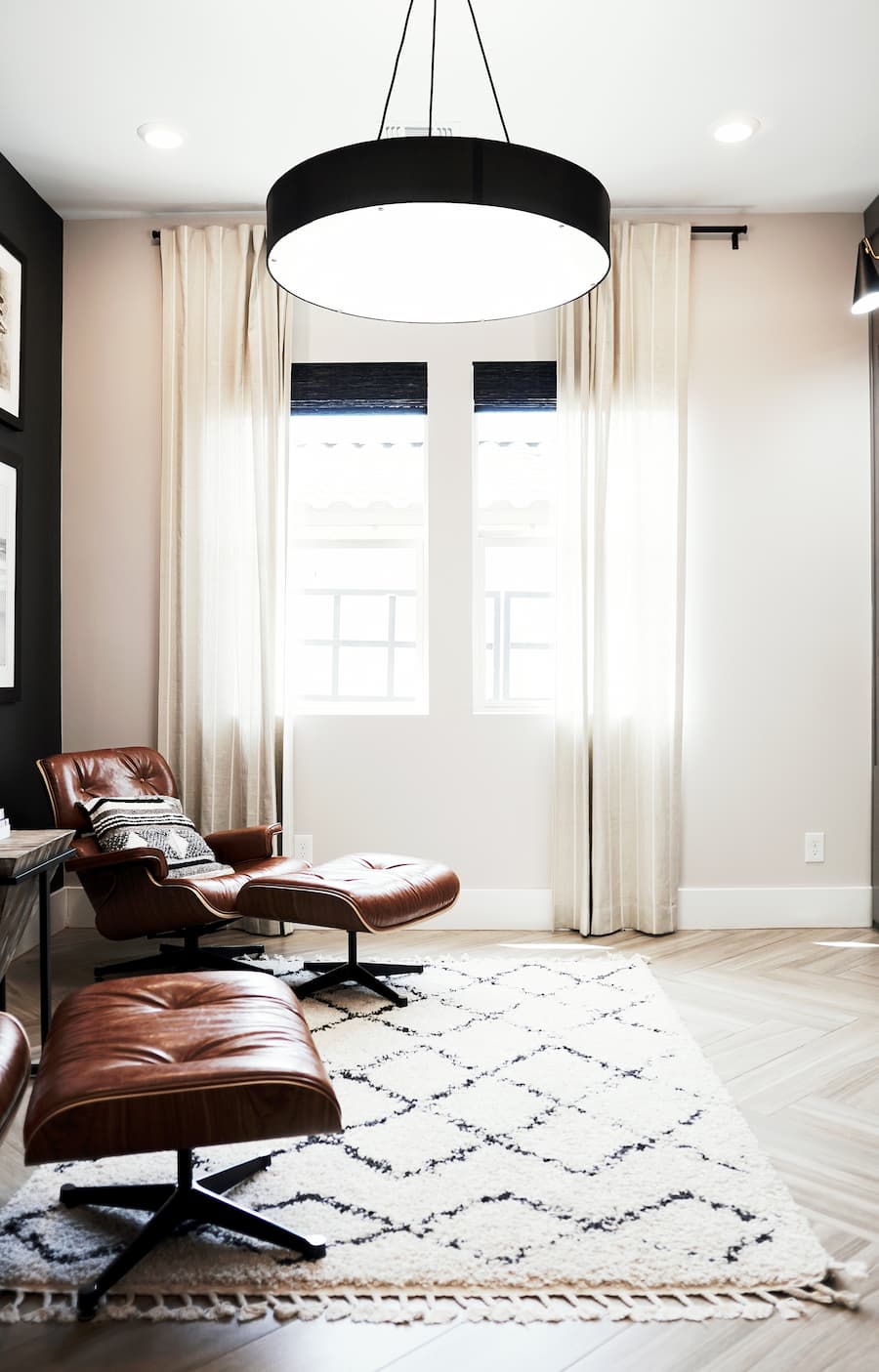Minimalist room painted in white with large black light pendant hanging from the ceiling. Brown leather Charles Eames chair and foot stool positioned to the left of the room against a wall painted in black and a cream rug positioned on the floor in the centre with a black diamond outline pattern