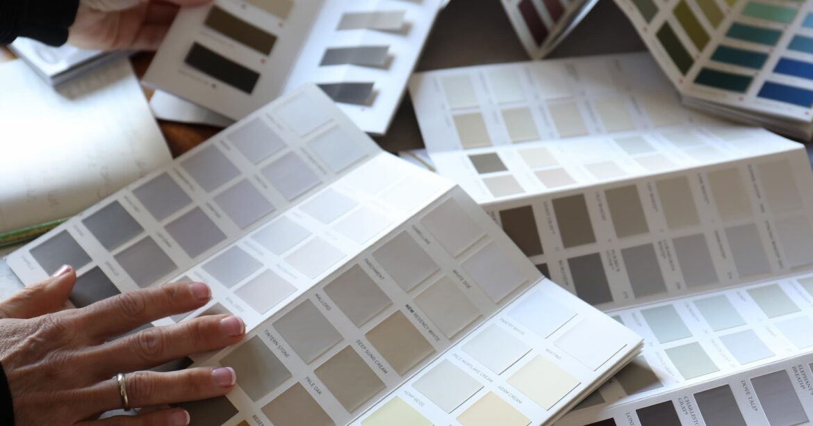 A person’s hands holding open a colour swatch card. The card is open on a page with different shades of grey. There are other colour swatch cards scattered around the table. The table is made of wood and there is a white notebook and laptop on the table.