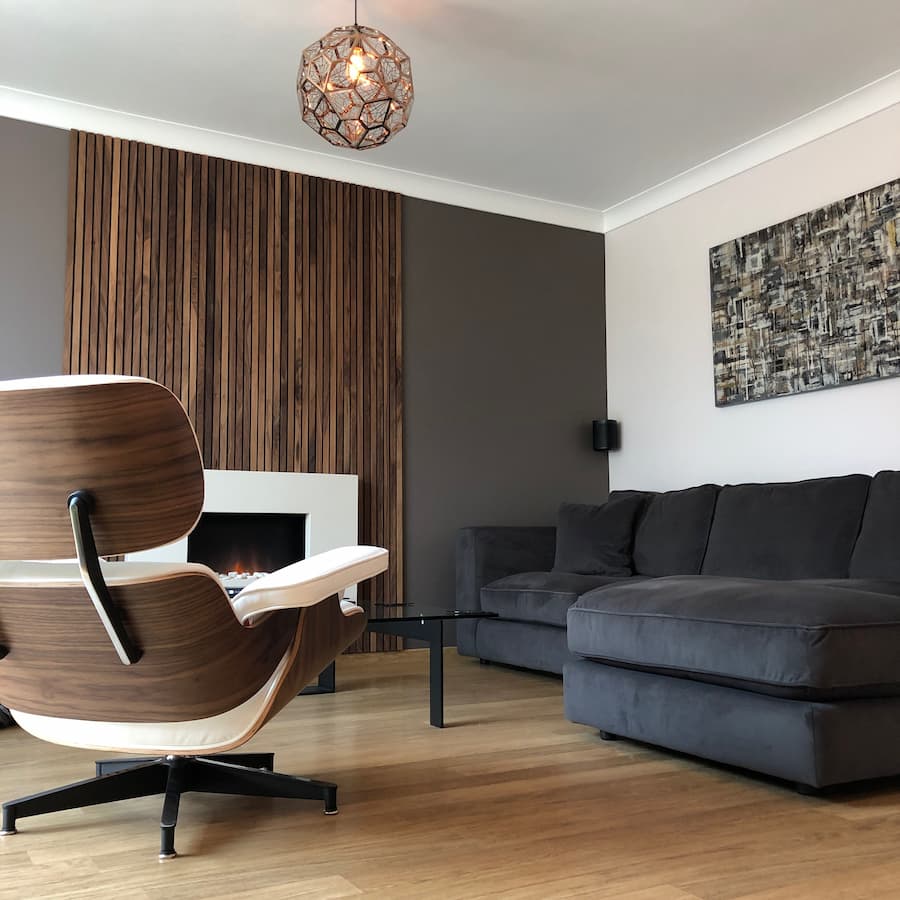 Living room with walnut panelled wall feature in the middle against a dark brown painted feature wall. Charles Eames chair in walnut and white leather positioned to the left and a dark grey corner sofa in velvet to the right