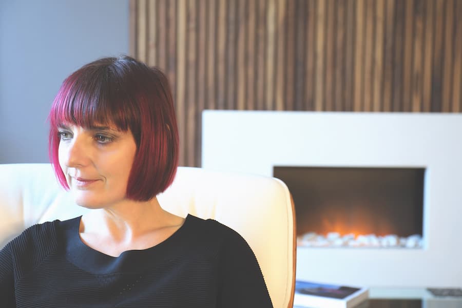 A person sitting on a white armchair in a living room. The person has short red hair and is wearing a black top. The background consists of a vertical wooden panelled feature wall and a white fireplace with a lit fire. The mood of the image is cozy and relaxed.