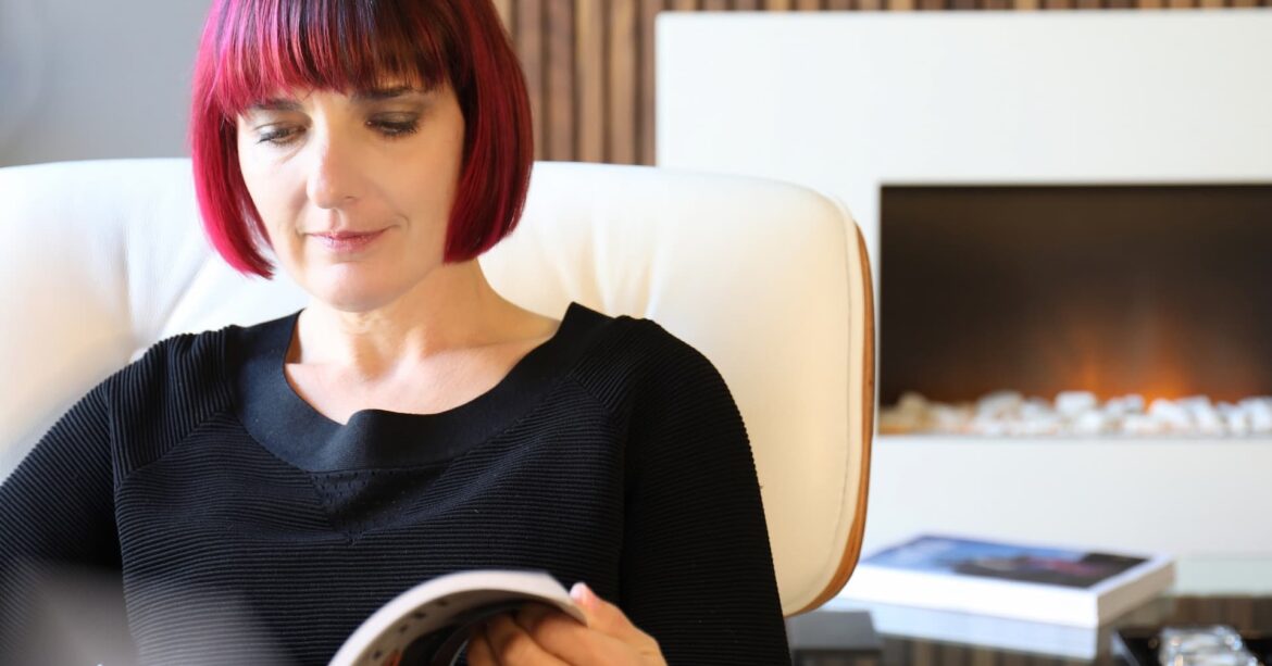 female with red hair sat in a white chair flicking through a magazine