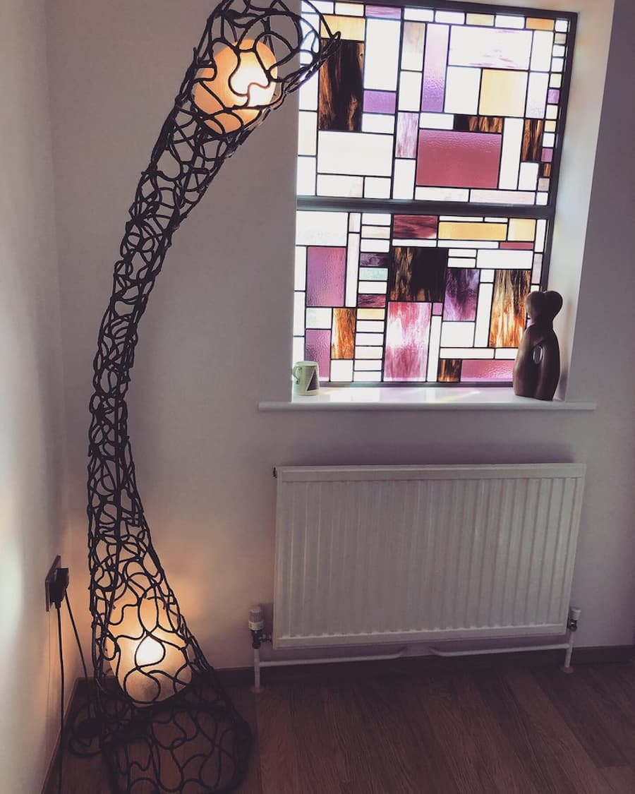 Corner of lounge with a tall, abstract lamp and a stained glass window. The lamp is made of brown leather and has a bulb at the top and bottom. It is curved and leans towards the window. The window is made of different colours and shapes of stained glass. The colours are mostly pink, purple, and brown.