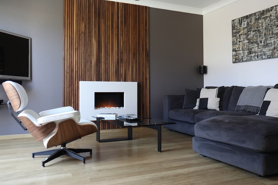 A modern living room with a fireplace and a wooden accent wall. The room has a wooden floor and a grey corner sofa with a throw and pillows. There is a white armchair with a wooden back and a coffee table with black legs and a glass top in front of the sofa. The fireplace is white and built into the wooden accent wall. There is a brown and white abstract painting hanging above the sofa.